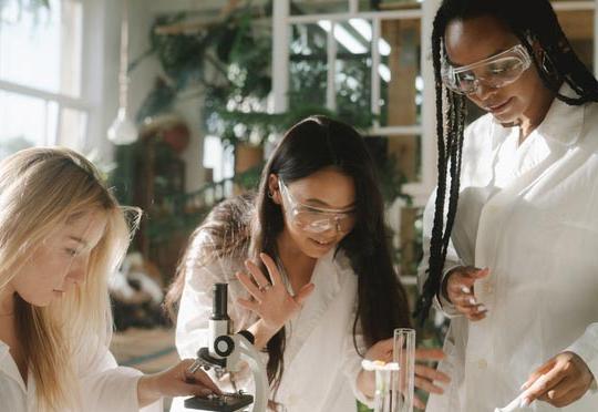 一个扎着辫子的黑人妇女的照片, 一名亚洲女子和一名金发女子戴着透明的安全护目镜，穿着白色的实验室大褂，挤在各种实验室设备周围 